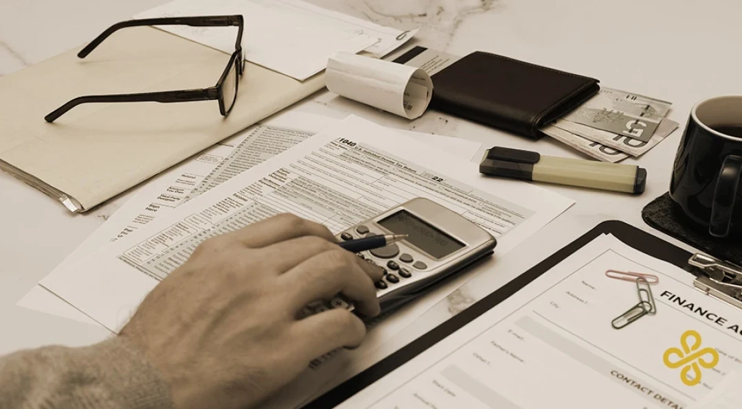 A hand using a calculator on a desk full of finance paperwork and other office equipment